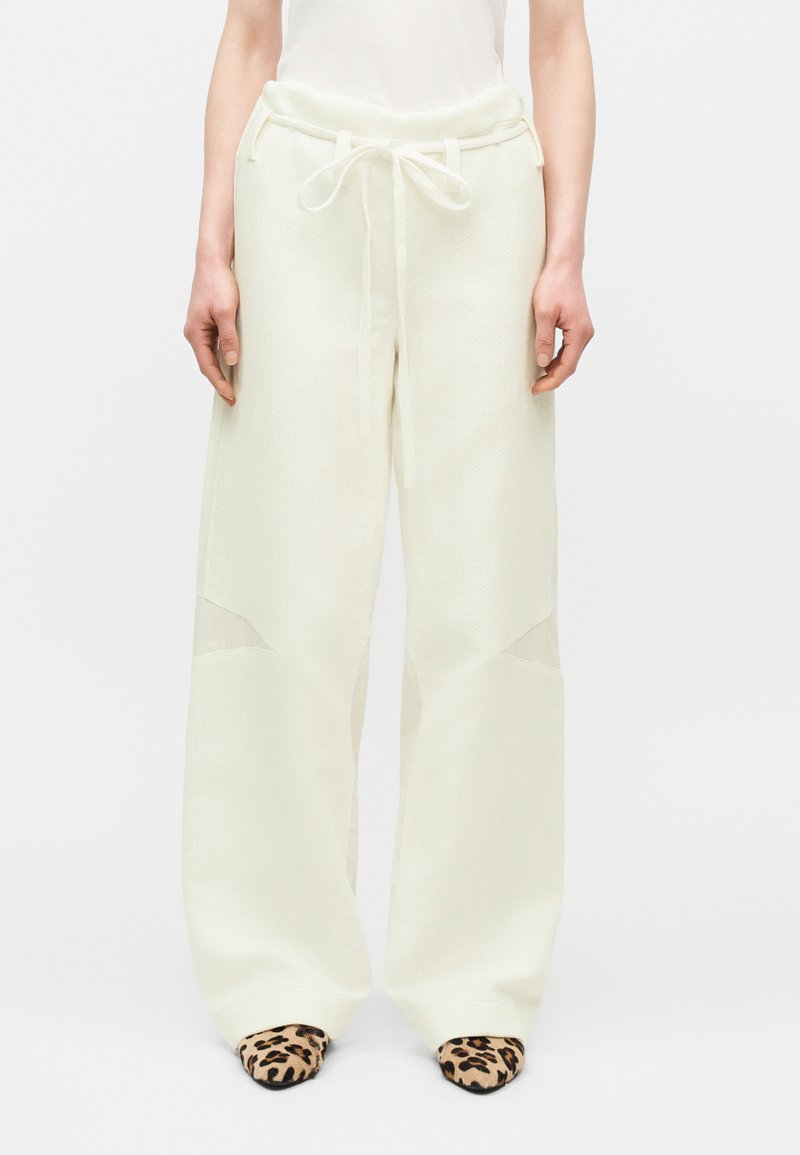 Personne portant un pantalon ample blanc texturé avec une taille nouée et des chaussures pointues imprimées léopard, debout devant un fond uni.
