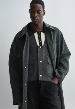 Sötétzöld gyapjú kabát nagy gallérral, egy szürke kötött mellény felett, amelynek közepén krém színű csík és gombok találhatóak, fekete farmerral párosítva.