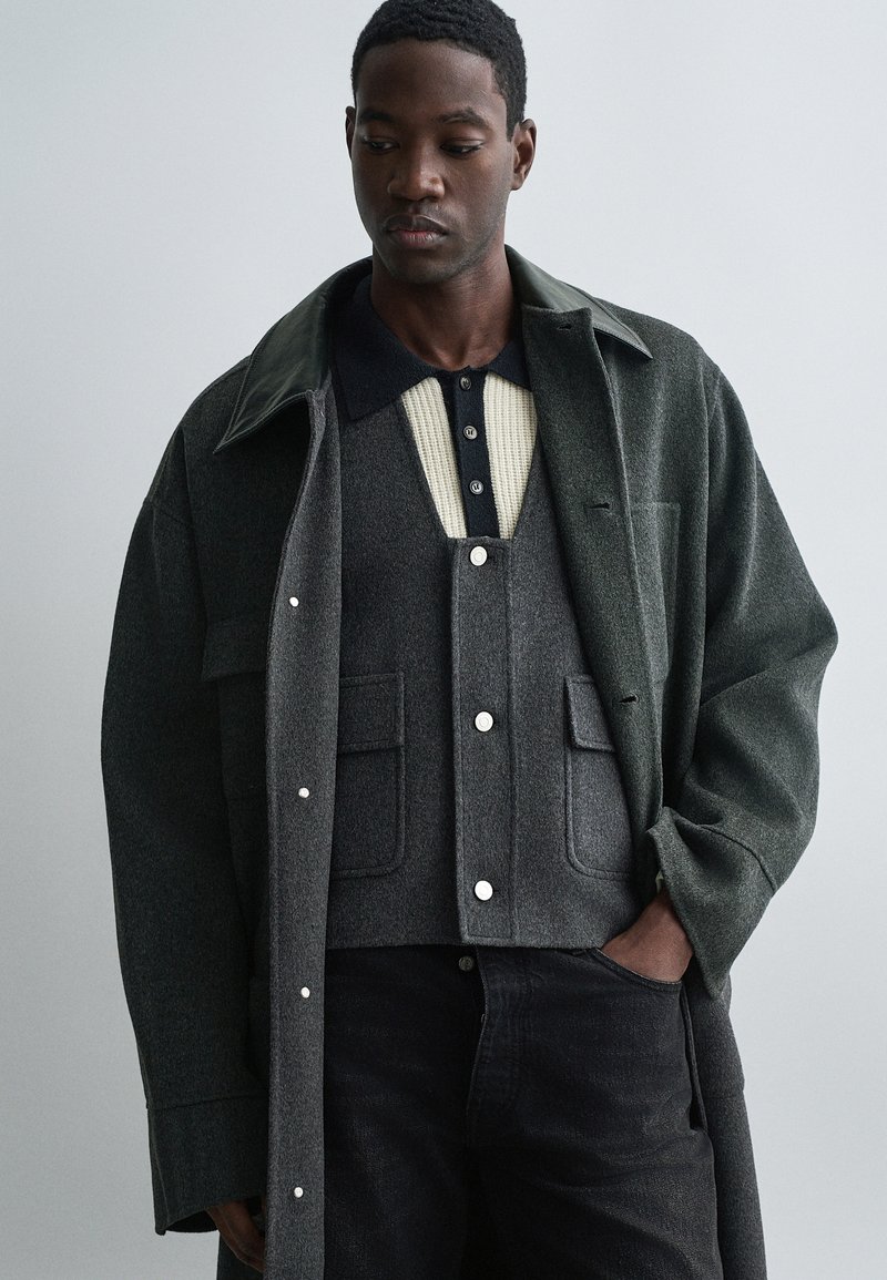 Tumši zaļš vilnas mētelis ar lielu apkakli, virs pelēka adīta veste ar krēmīgu centrālo svītru un pogām, kopā ar melniem džinsiem.