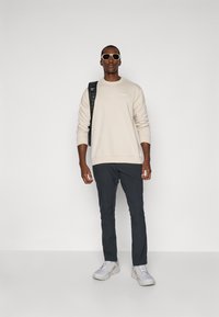 Sweatshirt bege claro com decote redondo, calças cinza escuro e ténis brancos. Acessórios incluem uma mochila e óculos de sol claros.
