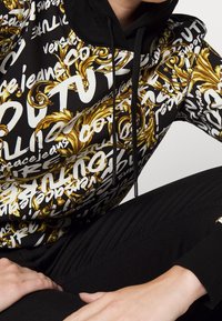Mikina s kapucí s výrazným černým pozadím, na kterém se nacházejí bílé a zlaté barokní textové vzory. Stahovací šňůrky a volný střih dotvářejí design.