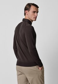 Dunkelbrauner Pullover mit Kragen, figurbetontem Schnitt, langen Ärmeln und geripptem Saum, kombiniert mit hellen Hosen.