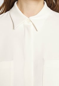 Camicia bianca con bottoni e colletto classico, due tasche sul petto e tessuto liscio e strutturato. Presenta una chiusura con bottoni anteriore.
