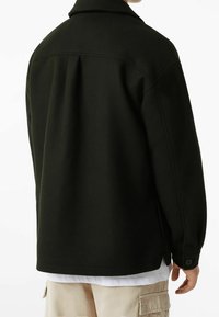 Černá bunda s límcem, vyrobená z měkkého materiálu. Má zadní pás a knoflíkové manžety. Nositá přes bílou košili a béžové kalhoty.