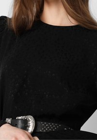 Femme portant un haut noir texturé à manches longues avec des épaules bouffantes et une large ceinture noire avec une boucle décorative argentée.