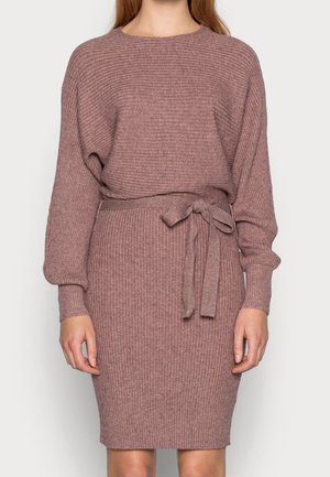 Femme portant une robe tricotée à manches longues côtelées avec un lien à la taille rose poudré, visible de la moitié du cou jusqu’au-dessus des genoux.