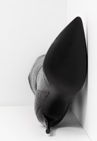 Zapato de tacón alto negro con textura, punta afilada y suela lisa, con un ligero patrón reptiliano en el material superior.