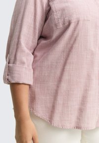 Nahaufnahme einer Person, die ein pinkes, kariertes Langarmhemd mit hochgekrempelten Ärmeln und weißen Hosen trägt.