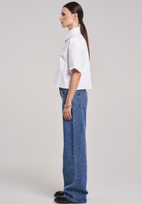 Cropped white button-up shirt with short sleeves, chest pocket, paired with wide-leg blue denim jeans and black shoes, viewed from the side.