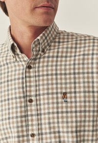 Cămașă cu carouri maro și alb, cu guler butonat și design brodat cu urs. Prezintă butoane maro și un material texturat.