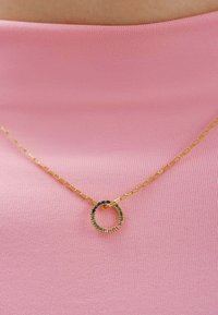 Collar de cadena de oro con un colgante circular adornado con piedras verdes, azules y moradas, sobre un fondo de tela rosa.