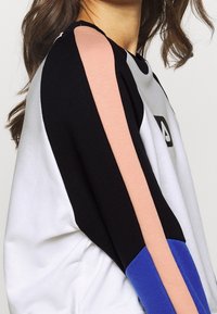 Sweatshirt coloré avec des sections noires, blanches, roses et bleues, présentant un col rond et un détail de logo bien visible à l'avant.