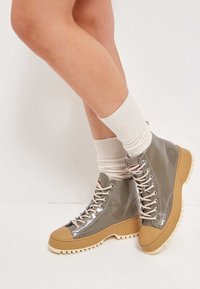 Buty za kostkę z błyszczącego szarego materiału z fakturowaną, beżowo-brązową gumową podeszwą, wyposażone w okrągłe metalowe oczka i beżowe sznurówki.