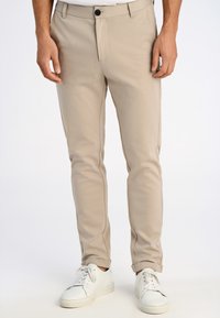 Man draagt beige slim-fit broek met omgeslagen zomen en witte veterschoenen, staand tegen een effen lichte achtergrond.