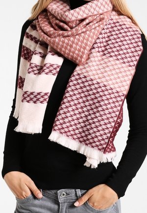 Personne portant une grande écharpe à motif pied-de-poule dans des tons de rose et de bordeaux sur un haut à manches longues noir, les mains dans les poches d'un jean gris.