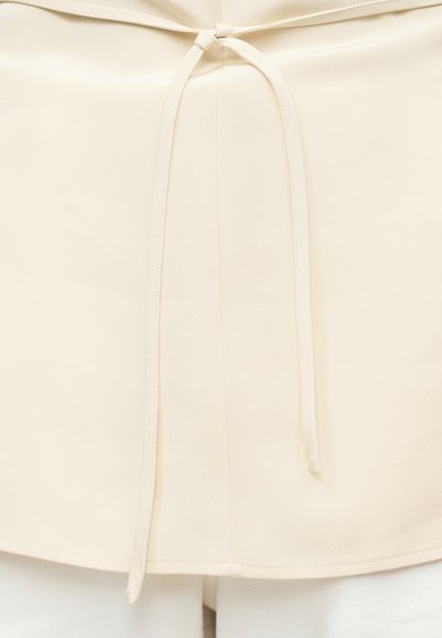 Corset ceinture en tissu crème nouée en un simple nœud à l'avant, portée sur des vêtements blancs.