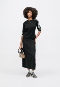 Čierny texturovaný sveter s bielymi pruhmi, v kombinácii s dlhou čiernou sukňou s bočnými rozparkami. Sivé športové topánky a priehľadná kabelka.