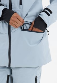 Hellblau wasserfeste Jacke mit schwarzen Akzenten. Verfügt über eine Reißverschlusstasche, elastische Bündchen und eine glatte, langlebige Stoffstruktur.