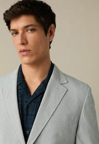 Blazer a righe grigio chiaro con rever a intaglio, chiusura a un bottone, tessuto texturizzato, indossato sopra una camicia blu scuro a fantasia.