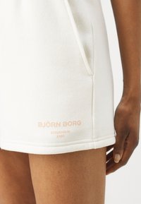 Abito in cotone bianco con una vestibilità relax, dotato di tasche laterali e logo ricamato in rosa pallido. Dettaglio di cucitura visibile all'orlo.
