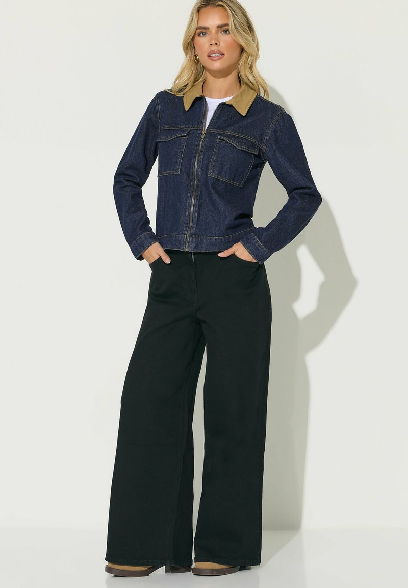 Donna che indossa una giacca di denim scuro con collo color cuoio, camicia bianca, pantaloni neri a gamba larga e scarpe marroni, in piedi con le mani in tasca.