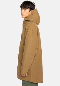 Junger Mann in einer beige Kapuzenjacke mit oversized Schnitt, der seitlich mit neutralem Gesichtsausdruck vor einem einfarbigen hellen Hintergrund steht.