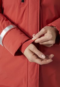 Chaqueta aislante roja con un tejido texturizado, que presenta una franja reflectante en el puño. Manos ajustando la apertura de la manga.