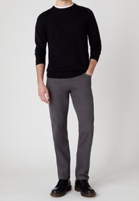 Man wearing black sweater, gray pants, and black leather shoes standing with one hand in pocket against plain background.