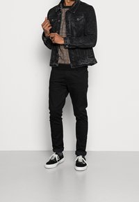 Giubbotto di jeans nero con due tasche sul petto, maglione a girocollo a fantasia, jeans neri e sneakers nere con suola bianca.