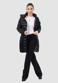 Piumino nero con chiusura a zip, cappuccio e design trapuntato. Indossato sopra una camicia bianca e pantaloni neri, con una vestibilità comoda.