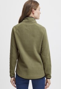 Pullover in pile verde oliva con colletto alto, maniche raglan e bordo nero all'orlo. Texture morbida, design minimal e senza hardware visibile.