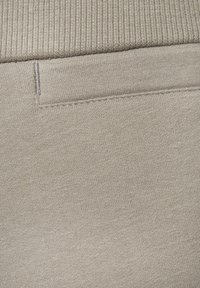 Beiger Stoff mit glatter Textur, ausgestattet mit einem horizontal gerippten Bund und einem dezenten gesteppten Taschen-Detail.