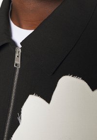 Chaqueta negra con un cuello estructurado y cremallera plateada. Presenta un gran diseño de aplique blanco con bordes deshilachados en la parte delantera.