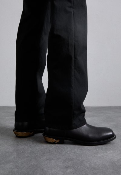 Svarta läderankelboots med rund tå, prydda med en dekorativ guldfärgad valdetalj på klacken. Bärs med svarta byxor.