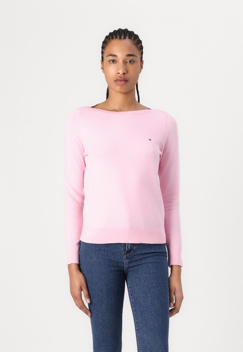 Lys rosa langermet genser med rund hals og en liten logo på brystet, sammen med mørkeblå jeans.