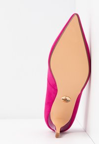 Chaussure à talon haut rose vif à bout pointu reposant sur le côté, montrant la semelle lisse couleur cuir clair et la petite pointe du talon sur fond blanc.