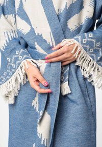 Modrý a biely vzorovaný záblesk s třásňovými rukávmi a viazaním zabezpečeným v páse. Materiál sa zdá byť mäkký s textúrovaným povrchom.