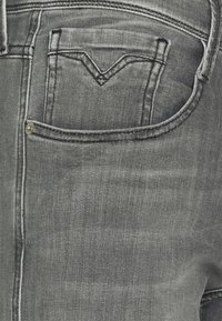 Jeans de mezclilla gris con un bolsillo trasero que presenta un patrón de costura en forma de V, botón estándar y una sutil variación en la textura.