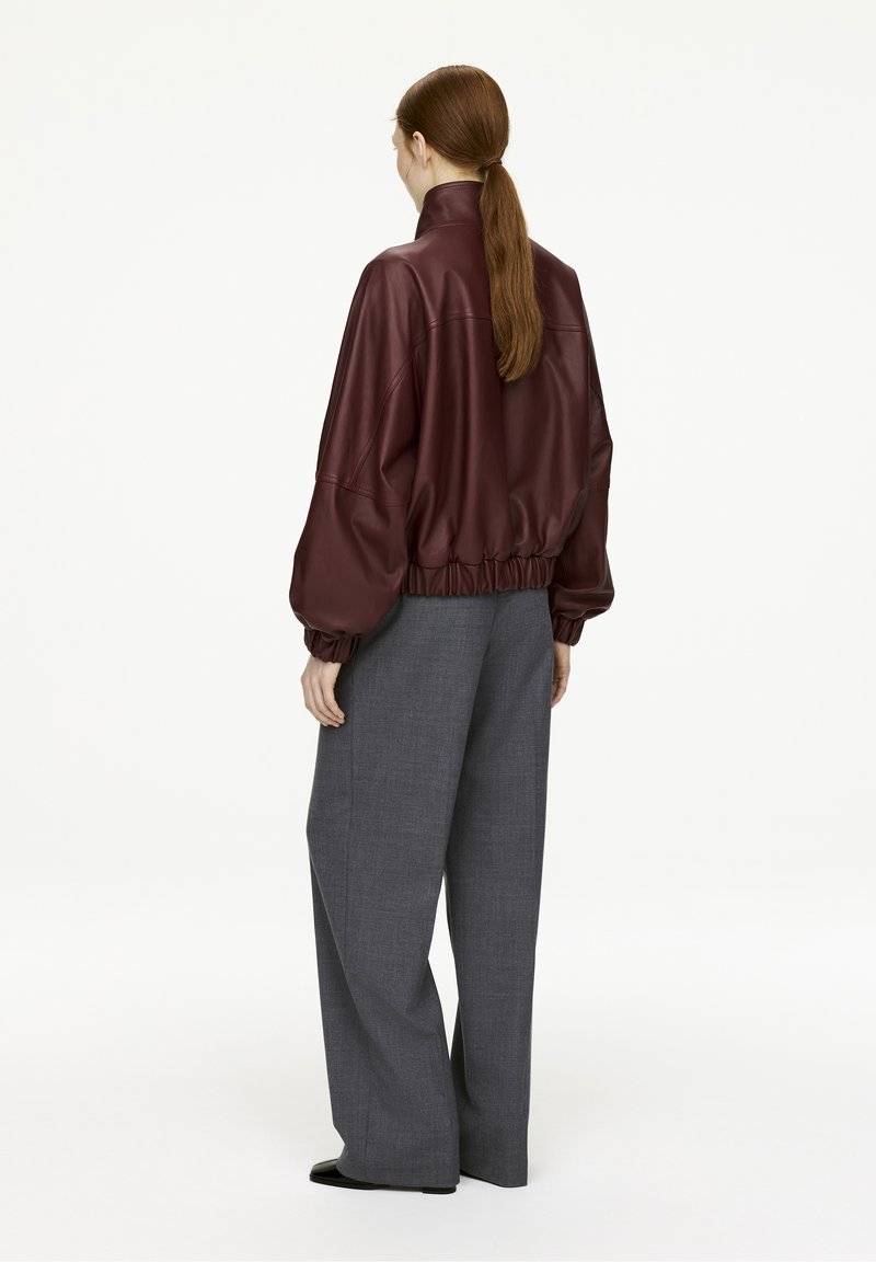 Burgundy leather jacket with a high collar, gathered hem, and oversized sleeves paired with gray wide-leg trousers and black footwear.