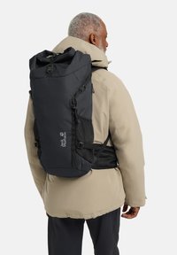 Zaino nero con design a rullo, spallacci regolabili, tasche laterali in rete e un logo. Indossato da una persona con una giacca beige.