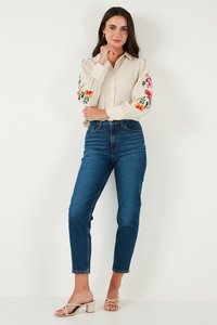 Camisa beige de botón con bordado floral en las mangas, combinada con jeans de tiro alto azul y sandalias de tacón beige con tiras.
