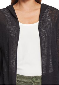 Černý, lehký pletený kardigan s kapucí, s průhlednou texturou a otevřeným předním dílem, vrstvený přes bílé tričko a zelené kalhoty.
