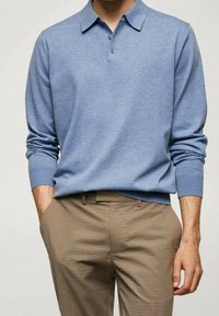 Uomo che indossa una polo azzurro chiaro a maniche lunghe infilata in pantaloni beige testurizzati, con le mani in tasca davanti a uno sfondo semplice.