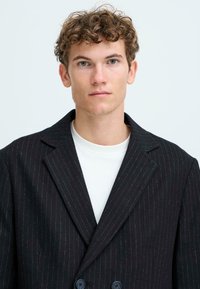 Black pinstriped blazer with a double-breasted design, wide lapels, and two dark buttons. Worn over a white crew-neck t-shirt.