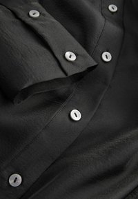 Must must, mis pehmeast kangast, valgete nuppudega äärisel ja volditud kraega. Tekstuur näib olema pehme ja sile.