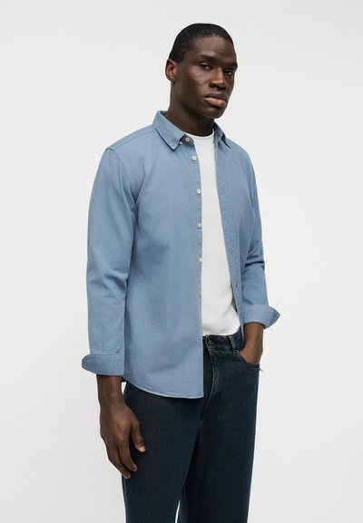 Ανοιχτό γαλάζιο πουκάμισο από βαμβάκι, με κλασικό γιακά, μακριά μανίκια που είναι γυρισμένα στις μανσέτες, και ένα λευκό T-shirt από κάτω.