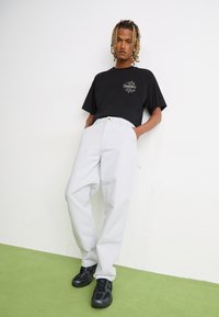 Zwarte oversized t-shirt met een grafisch ontwerp op de voorkant, gecombineerd met lichtgrijze baggy broek en zwarte sneakers. Soepele stof en ontspannen pasvorm.