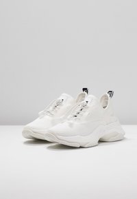 Scarpe sportive bianche realizzate in materiale a maglia traspirante, con suola spessa e sistema di lacci elasticizzati per una vestibilità sicura. Design minimalista.