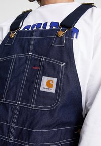 Tummansininen farkkuhaalari, jossa valkoiset ompeleet ja Carhartt-logolla varustettu kangasmerkki. Siinä on metalliset klipsit ja väljä istuvuus.