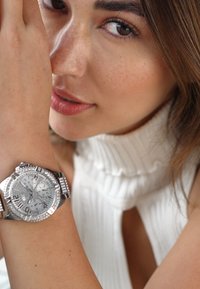 Montre bracelet en acier argenté avec un cadran texturé, des fonctions chronographe et des accents semblables à des diamants sur la lunette, portée sur un bras à la peau claire.